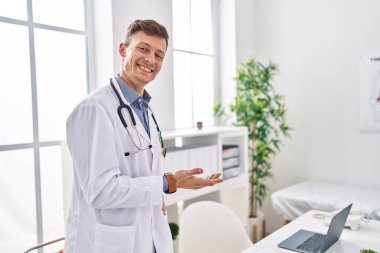 Young man doctor smiling confident standing with welcome gesture at clinic