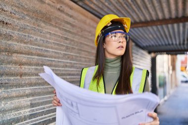 Young beautiful hispanic woman architect looking plans with serious expression at street