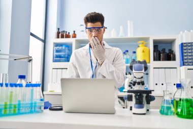 Young hispanic man working at scientist laboratory doing video call covering mouth with hand, shocked and afraid for mistake. surprised expression 