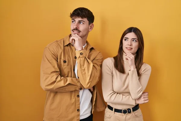 黄色の背景に立っている若いヒスパニック系のカップルは 質問を心配し 心配し 顎に手で緊張 — ストック写真