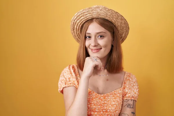 黄色い背景の上に立つ若い赤毛の女性は カメラを見て自信を持って見て夏の帽子をかぶって交差腕と手であごに提起笑顔 ポジティブな — ストック写真