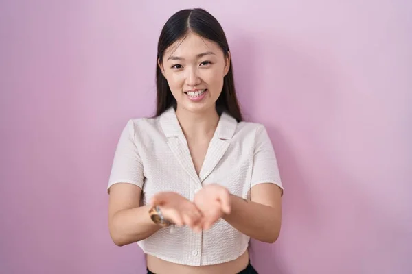 ピンクの背景の上に立つ中国の若い女性が一緒に手の手のひらで笑顔を受信またはジェスチャーを与える 保持し保護する — ストック写真