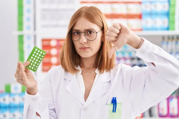 Young redhead woman working at pharmacy drugstore holding birth control pills with angry face, negative sign showing dislike with thumbs down, rejection concept