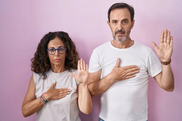 Középkorú Spanyol Házaspár Együtt Rózsaszín Háttér Esküszik Kéz Mellkason Nyitott — Stock Fotó