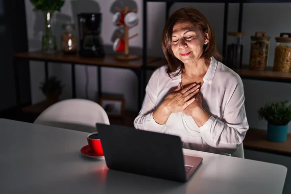 中年惊慌失措的女人晚上在家里用笔记本电脑微笑着 双手放在胸前 闭着眼睛 脸上挂着感激的手势 健康概念 — 图库照片