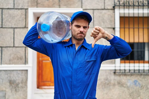 Young hispanic man holding a gallon bottle of water for delivery with angry face, negative sign showing dislike with thumbs down, rejection concept