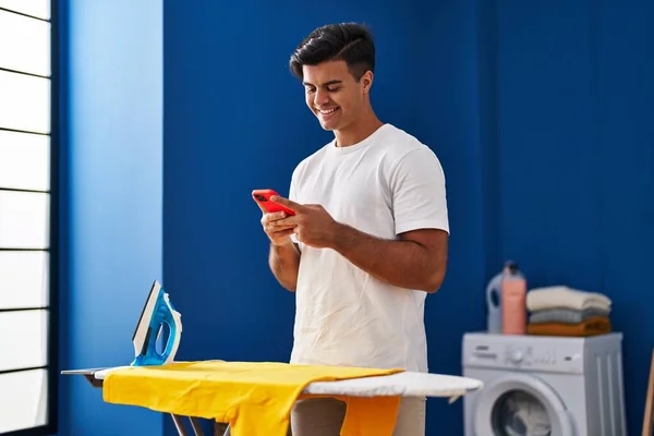 在洗衣房用智能手机熨烫衣服的年轻人 — 图库照片