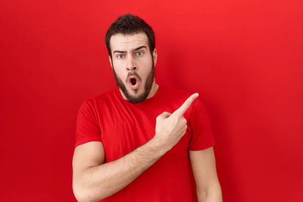 若いヒスパニック男を着てカジュアルな赤いTシャツ驚きを指して指で側に 口を開けて驚くような式 — ストック写真
