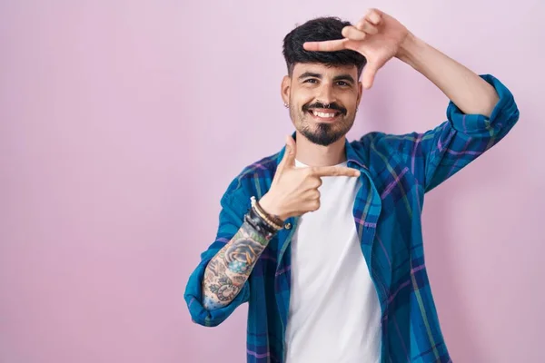 ひげを生やした若いヒスパニック系の男性がピンクの背景に立って 幸せな顔をした手と指でフレームを作って笑っています 創造性と写真の概念 — ストック写真