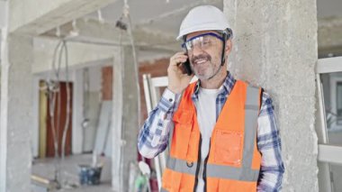 İnşaat alanında akıllı telefondan konuşan orta yaşlı bir inşaatçı gülümsüyor.
