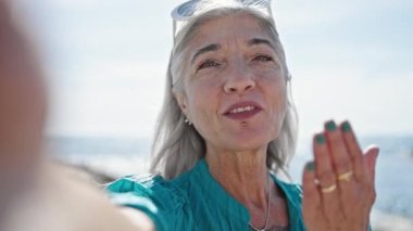 Orta yaşlı, kır saçlı bir turist kumsalda öpüşürken kamerayla selfie çekiyor.