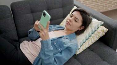 Beyaz kadın akıllı telefon kullanıyor. Evdeki kanepede yatıyor.