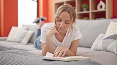 Young blonde woman reading book lying on sofa at home
