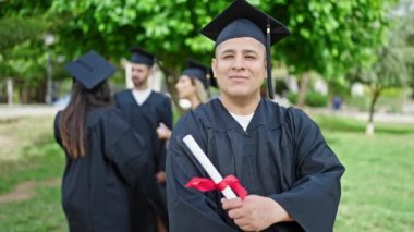 Bir grup öğrenci üniversite kampüsünde kollarını kavuşturarak diploma alarak mezun oldular.