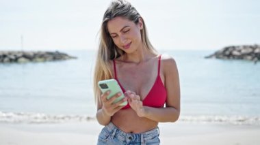 Kumsalda akıllı telefon kullanarak bikini giyen genç sarışın kadın turist.