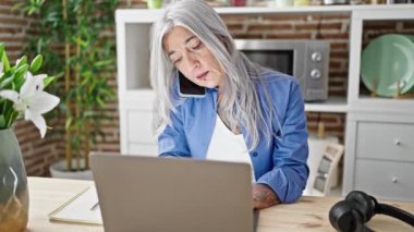Orta yaşlı, gri saçlı, dizüstü bilgisayar kullanan, yemek masasında oturan, akıllı telefonlu bir kadın.