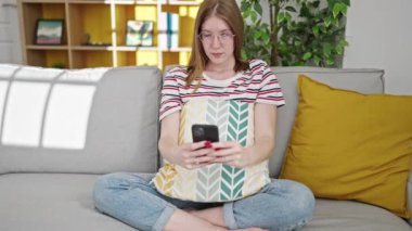 Evdeki akıllı telefonu kullanarak ciddi bir ifadeyle kanepede oturan genç sarışın kadın.