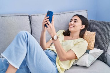 Evdeki kanepede uzanmış akıllı telefon kullanan genç İspanyol kadın.