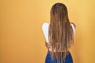 Sarı arka planda örülü saçları olan Afrikalı Amerikalı bir kadın mutlu ve pozitif bir şekilde kendine sarılıyor. Sevgi ve özgüven. 