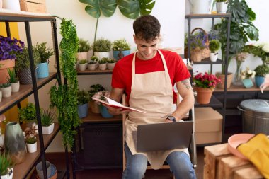 Young hispanic man florist using laptop reading book at flower shop