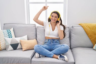 Young hispanic woman dancing and listening to music sitting on sofa at home