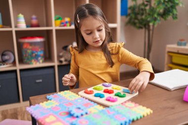 Adorable hispanic girl playing with maths puzzle game sitting on table at kindergarten