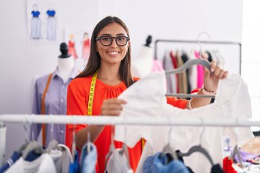 Young beautiful hispanic woman tailor smiling confident holding t shirt on rack at tailor shop