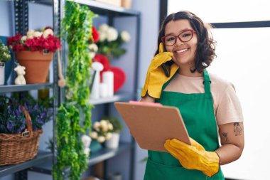 Young woman florist talking on smartphone reading clipboard at florist