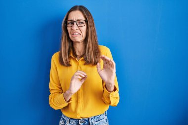 Mavi arka planda duran gözlüklü genç bir kadın tiksinti reaksiyonu yüzünden tiksinmiş ve ürkek bir ifade takınıyor. Eller havada. 