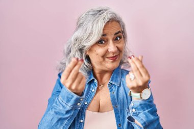 Middle age woman with grey hair standing over pink background doing money gesture with hands, asking for salary payment, millionaire business 
