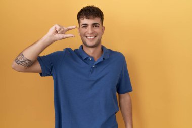 Young hispanic man standing over yellow background smiling and confident gesturing with hand doing small size sign with fingers looking and the camera. measure concept. 