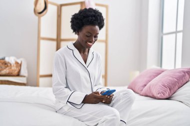 Young african american woman using smartphone sitting on bed at bedroom