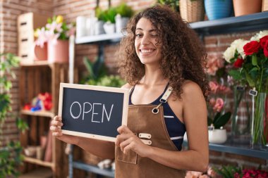 Çiçekçide çalışan kıvırcık saçlı İspanyol kadın elinde açık tabela, kameraya seksi bir ifadeyle bakıyor, neşeli ve mutlu bir yüzle.. 