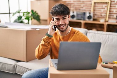 Young hispanic man using laptop talking on smartphone at new home