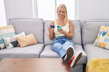 Akıllı telefon ve kredi kartı kullanan genç sarışın kadın evdeki koltukta oturuyor.