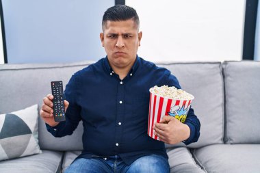 Latin asıllı genç adam TV 'de patlamış mısır yiyor şüpheci ve gergin, sorun yüzünden surat asıyor. negatif kişi. 