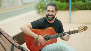 Young hispanic man musician playing classical guitar sitting on bench at park