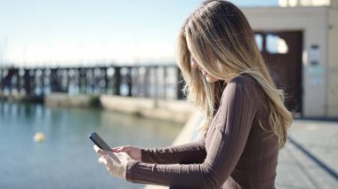 Young blonde woman smiling confident using smartphone at seaside