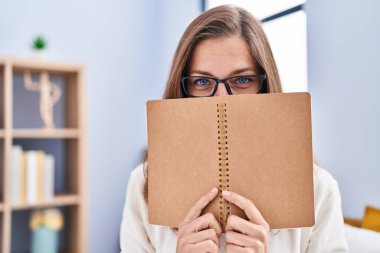Young woman covering face with book sitting on bed at bedroom