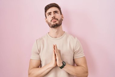 Hispanic man with beard standing over pink background begging and praying with hands together with hope expression on face very emotional and worried. begging. 