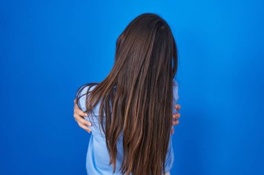 Genç esmer kadın mavi arka planda dikilip kendine sarılıyor mutlu ve pozitif bir şekilde. Sevgi ve özgüven. 