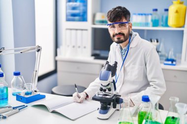 Young hispanic man scientist writing on notebook working at laboratory