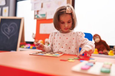 Adorable hispanic girl playing with maths puzzle game sitting on table at kindergarten