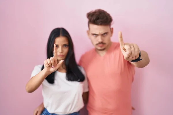 stock image Young hispanic couple standing over pink background pointing with finger up and angry expression, showing no gesture 