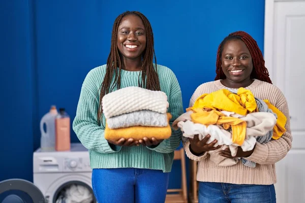 Two african women holding folded laundry after ironing smiling with a happy and cool smile on face. showing teeth.