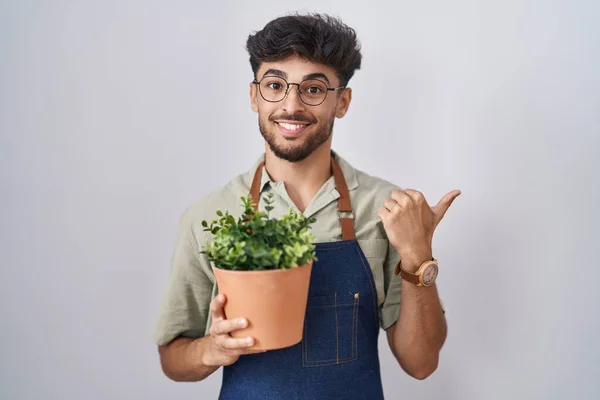 一个阿拉伯男人 留着胡子 手里拿着绿色的花盆 手拿着大拇指 背对着后面 带着自信的微笑 — 图库照片
