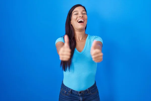 青い背景の上に立つ若いヒスパニック系の女性は 手で積極的なジェスチャーを行うことを承認します 親指アップ笑顔と成功のために幸せ 勝者のジェスチャー — ストック写真