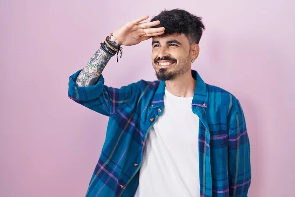 頭の上に手で遠くを見て非常に幸せと笑顔ピンクの背景の上に立ってひげを持つ若いヒスパニック系の男 検索の概念 — ストック写真