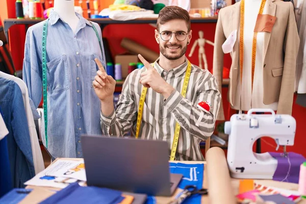 一个留着胡子的西班牙男人在裁缝店用笔记本电脑微笑着看着摄像机 用双手和手指指向旁边 — 图库照片