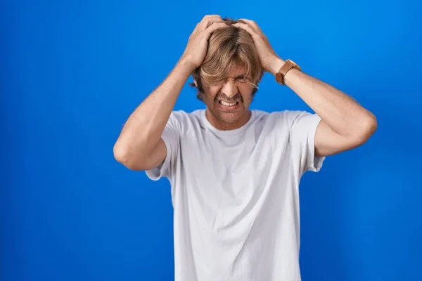 中年男人站在蓝色的背景上 由于疼痛和偏头痛 压力重重 手放在头上 — 图库照片